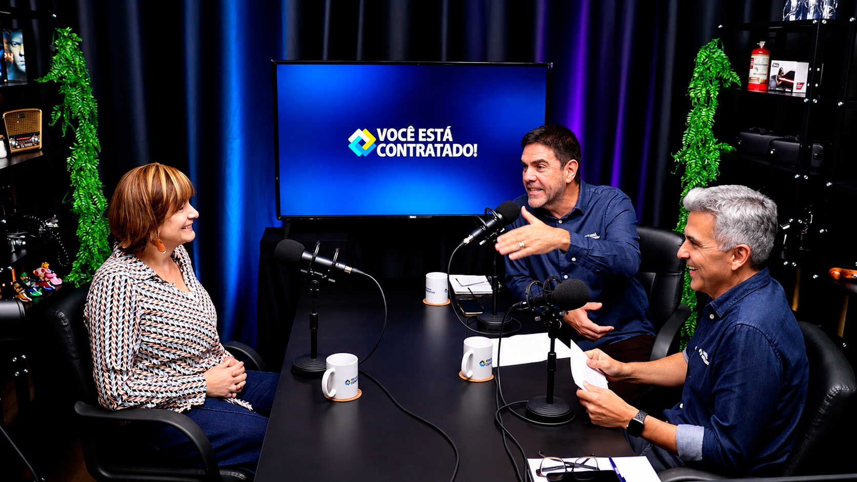 Três participantes conversam diante dos microfones durante a gravação de um podcast