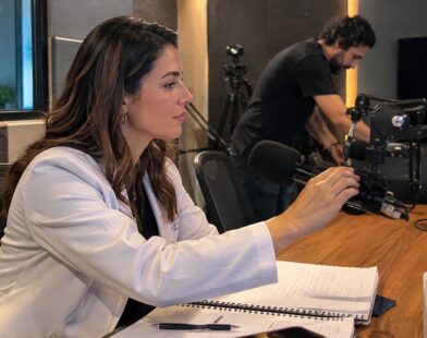 Médica prepara o microfone em estúdio de podcast enquanto técnico ajusta equipamentos ao fundo.