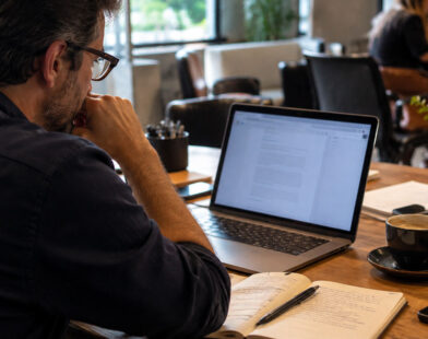 Profissional revisa um artigo no notebook com caderno e café em uma mesa de coworking.