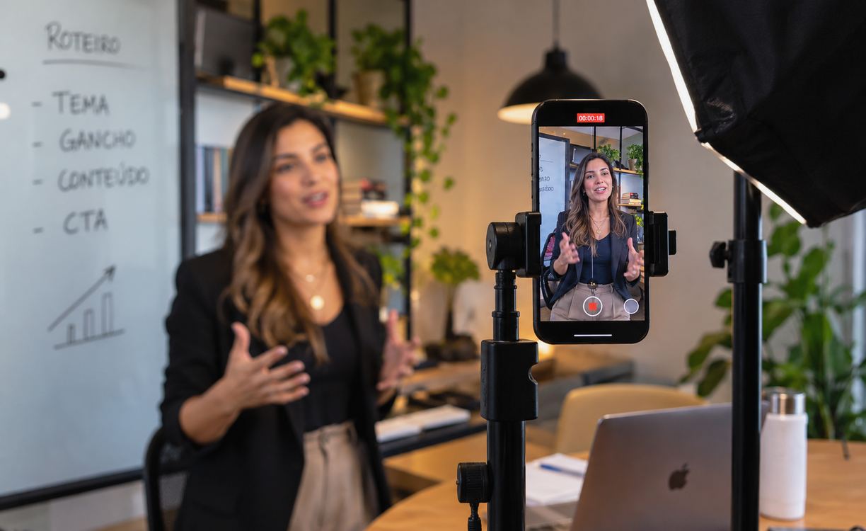 Celular em tripé grava apresentação vertical de profissional em ambiente de escritório, com iluminação e roteiro ao fundo.