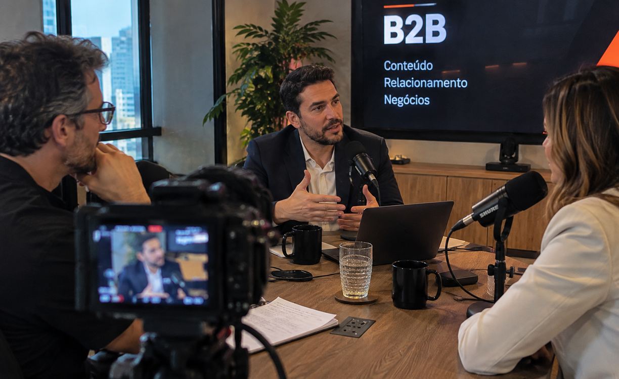 Três profissionais participam de uma gravação de podcast sobre negócios em uma sala de reuniões, com câmeras e microfones.