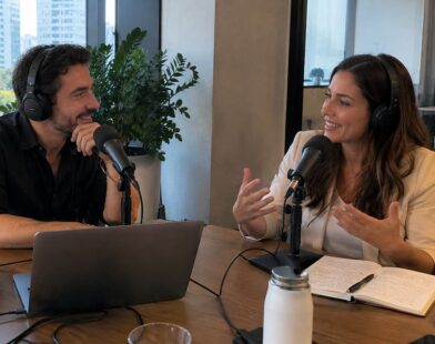 Dois profissionais conversam diante de microfones durante gravação de podcast em escritório, com notebook, anotações e ambiente corporativo.