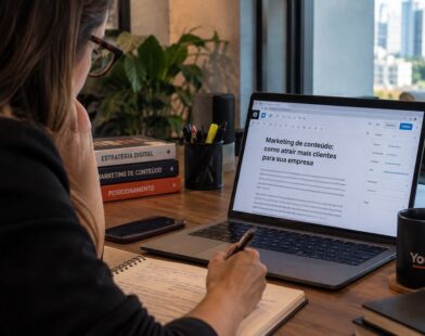Profissional revisa um artigo no notebook e faz anotações à mão em mesa de trabalho próxima à janela.